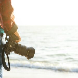 A person holding a DSLR camera with the ocean in the background at the beach.