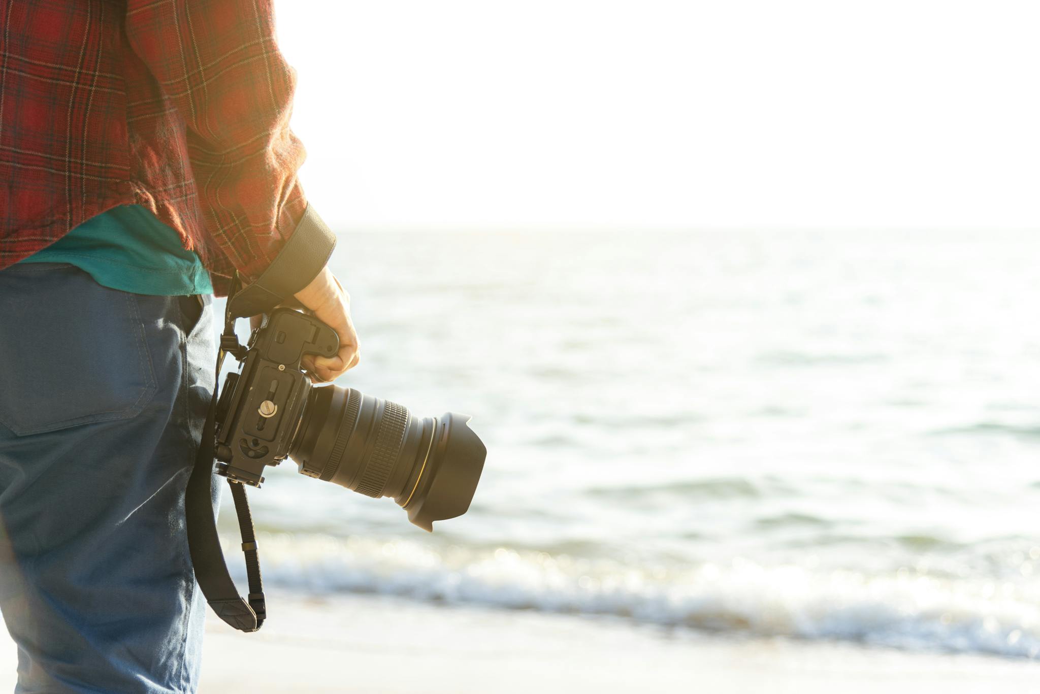 A person holding a DSLR camera with the ocean in the background at the beach.